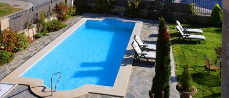 Une piscine rectangulaire bordée d’une terrasse en pierre, entourée d’un petit muret en pierre et d’une clôture en métal. On y trouve des chaises longues et un pot de fleurs près de la piscine.