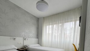 A hotel room with two beds, a bedside table with a lamp, a chair, and a window with sheer curtains. - IRAIPE GORBEA HOTEL (Vitoria-Gasteiz)