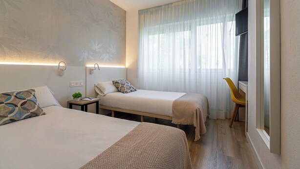 A hotel room with two beds, a small table, a chair, a TV, and a window with curtains. - IRAIPE GORBEA HOTEL (Vitoria-Gasteiz)