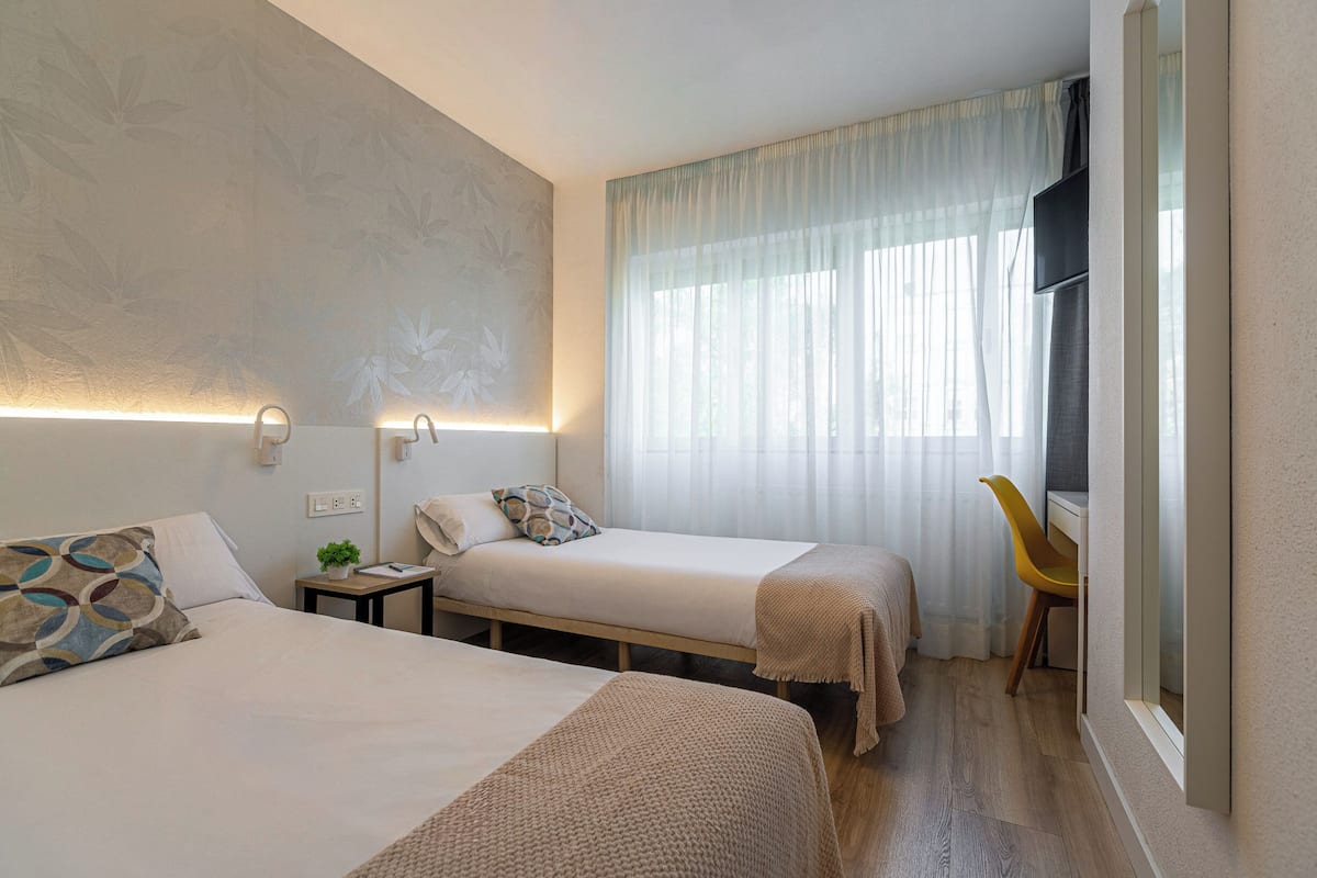 A hotel room with two beds, a small table, a chair, a TV, and a window with curtains.