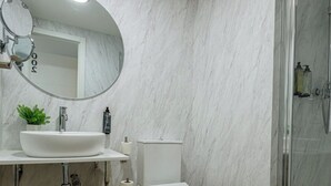 A modern bathroom with a marble wall, a round mirror, a white sink, a toilet, and a glass shower enclosure. - IRAIPE GORBEA HOTEL (Vitoria-Gasteiz)