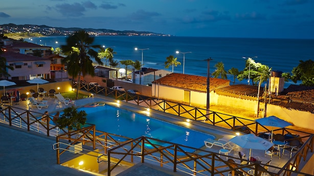 Une piscine avec vue sur l’océan, entourée de palmiers et bordée d’un bâtiment dont le toit est recouvert de tuiles.