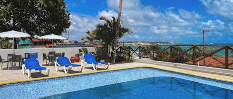 A pool area with blue tiled flooring, white lounge chairs, and a palm tree.