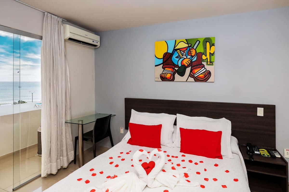A hotel room with a bed, red pillows, a painting on the wall, a glass table, and a view of the sea.
