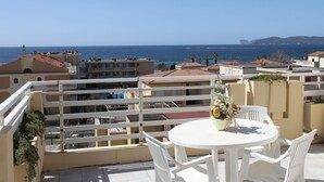 En balkon med hvidt bord og stole, med udsigt over et kystområde med bygninger og havet.