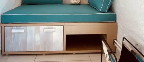 A small, cozy room with a teal bed, a window with curtains, a wall-mounted light fixture, and a small shelf with a decorative item.