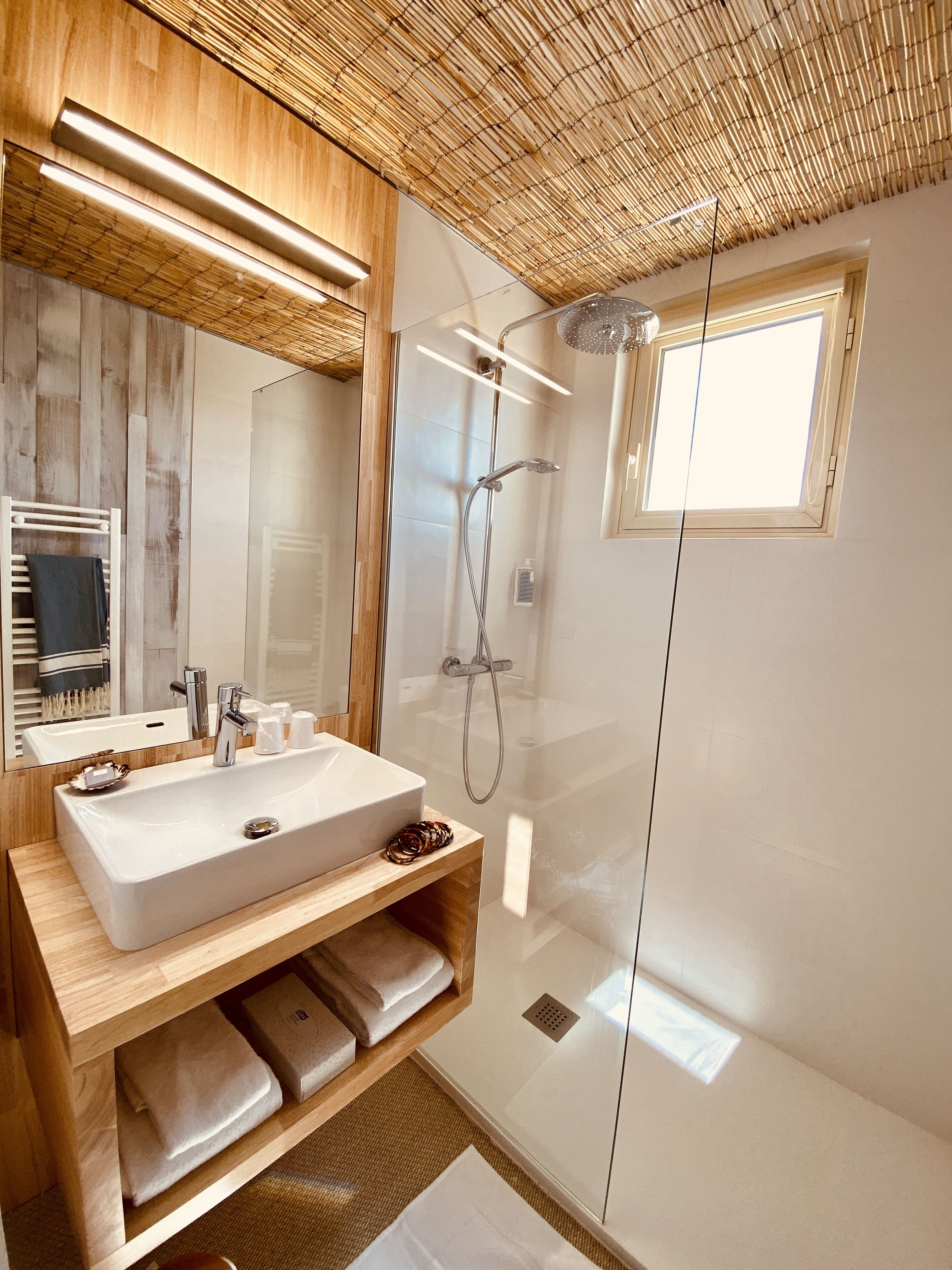 A modern bathroom with a large mirror, a wooden vanity, a glass-enclosed shower, and a small window.
