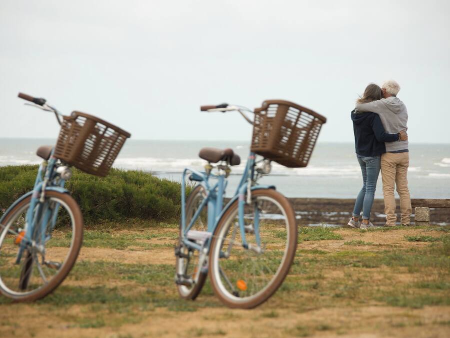 Dos bicicletas con cestas, una pareja de pie junto al océano y un banco de madera.