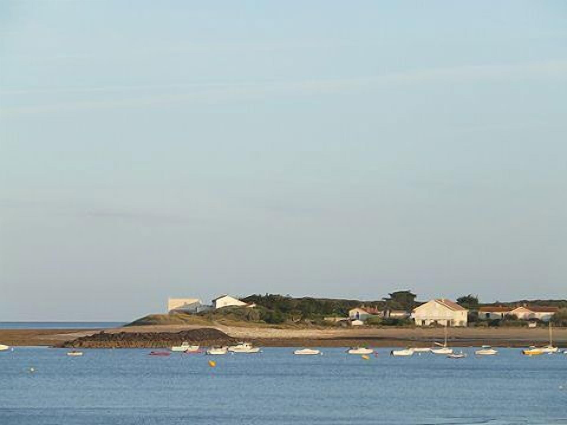 A coastal scene with several boats anchored in the water, a sandy beach, and a row of houses with a hill in the background.