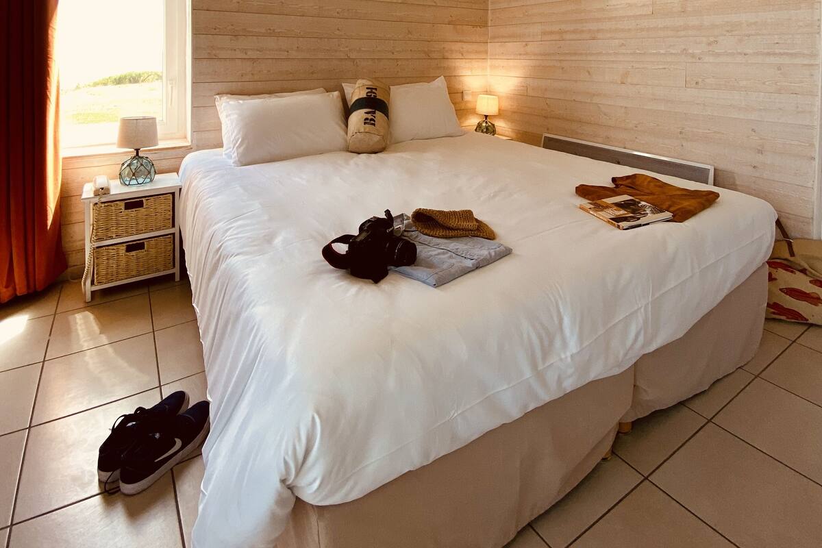 Une chambre avec un grand lit, des murs en bois, une fenêtre avec des rideaux, une lampe de chevet et une table de chevet avec une lampe et un appareil photo.