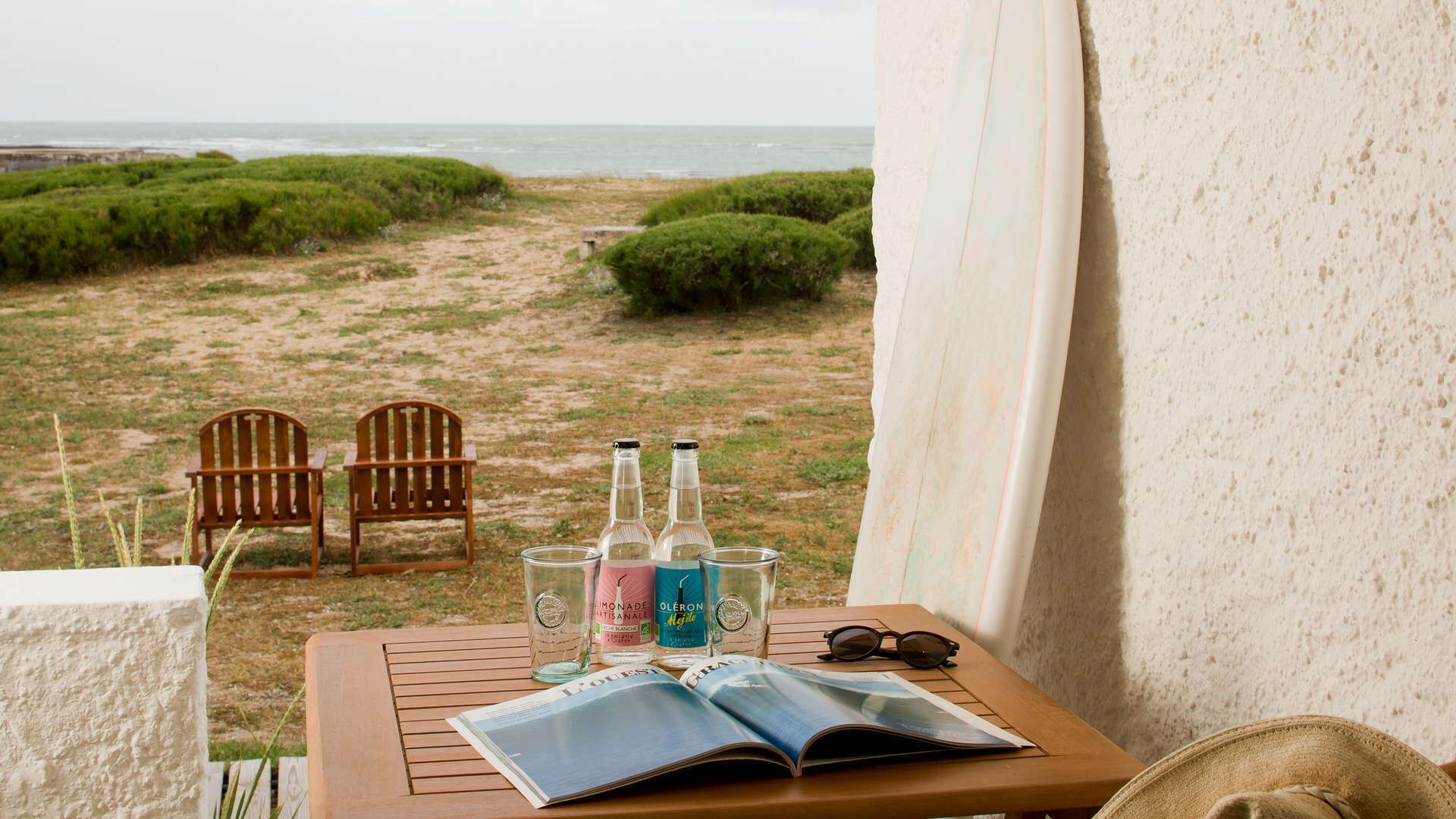 Un balcón con vista a la playa, dos sillas de madera, una mesa con botellas y una revista, y un par de gafas de sol.