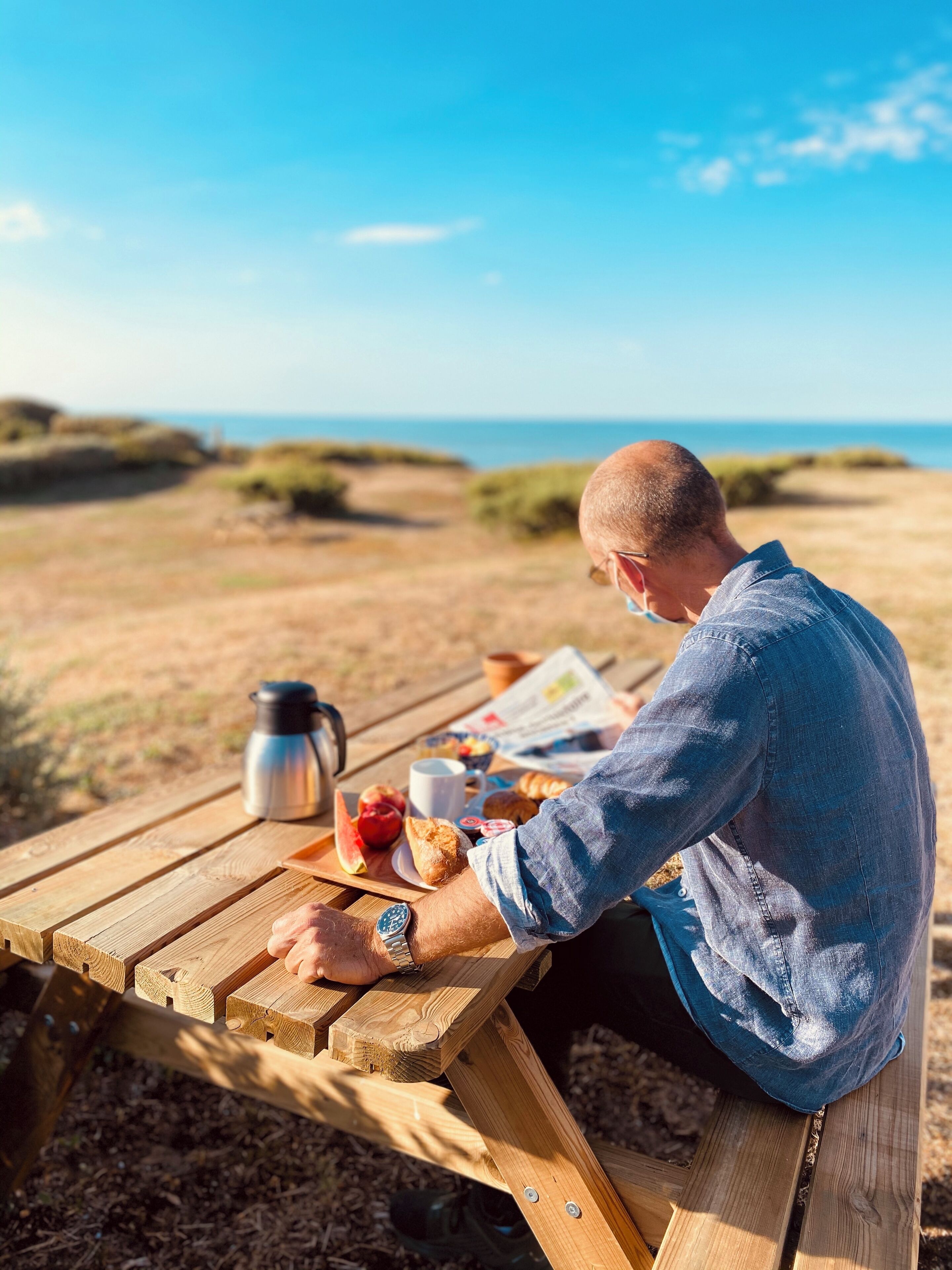 A person sitting at a wooden picnic table with a meal, a thermos, and a newspaper.