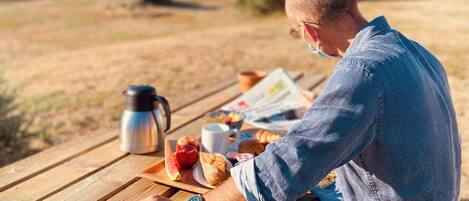 A person sitting at a wooden picnic table with a meal, a thermos, and a newspaper.