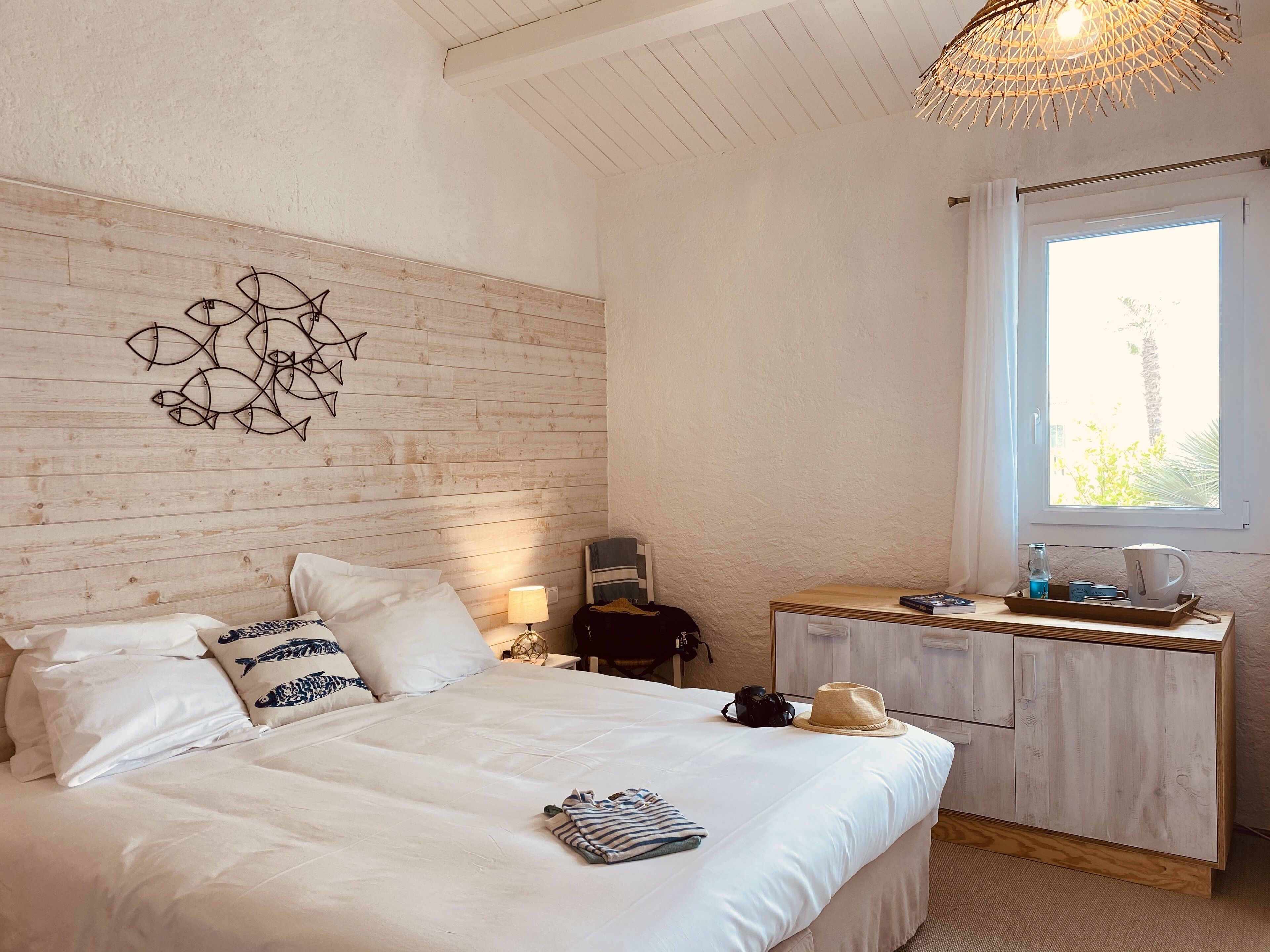Ein Schlafzimmer mit Holz-Kopfteil, einem Bett mit weißer Bettwäsche, einem Nachttisch mit Lampe, einem Schminktisch mit Spiegel und einem Fenster mit Blick auf Palmen.