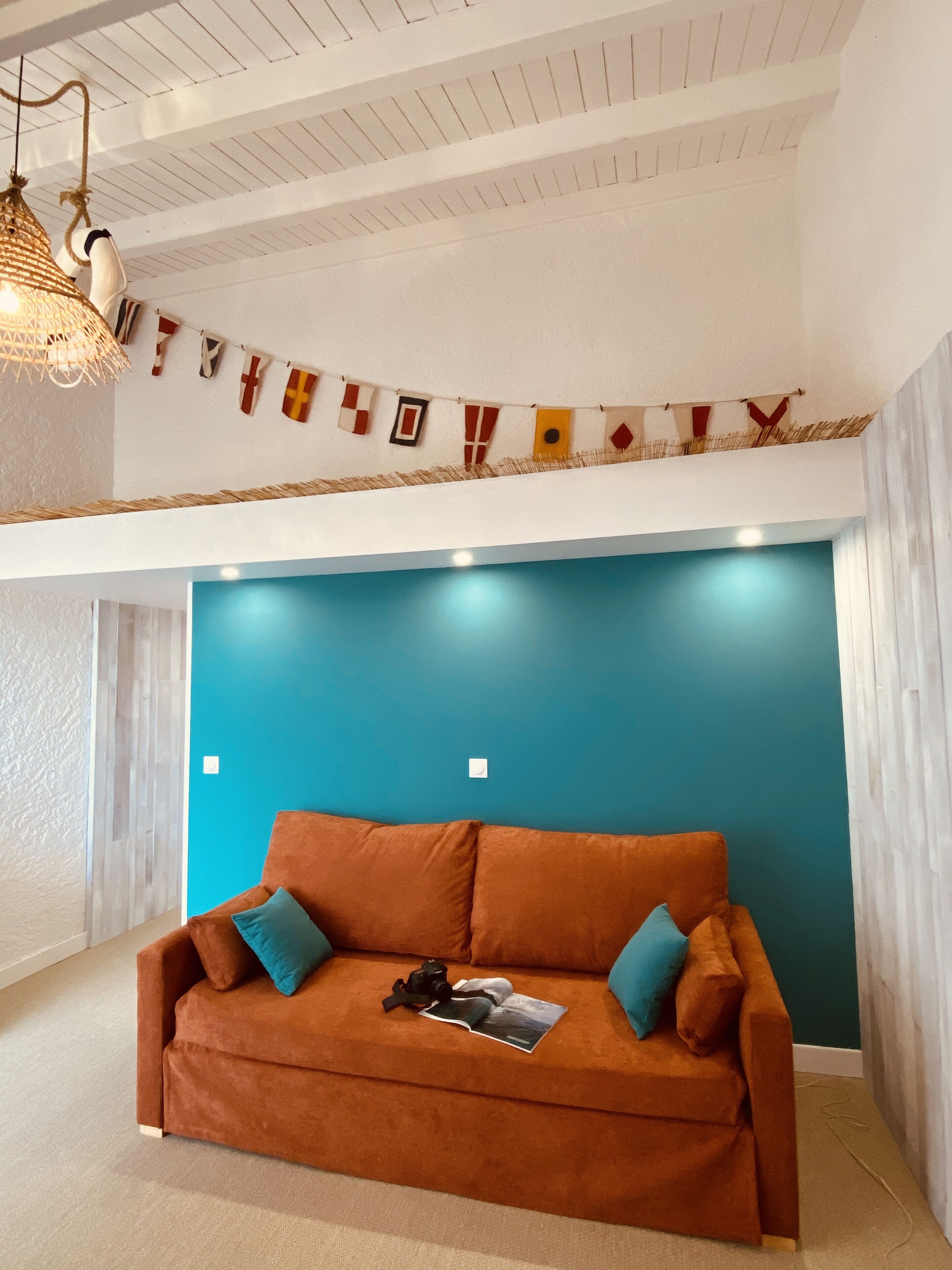 A cozy room with a blue accent wall, a brown sofa with cushions, a hanging light fixture, and a string of flags.