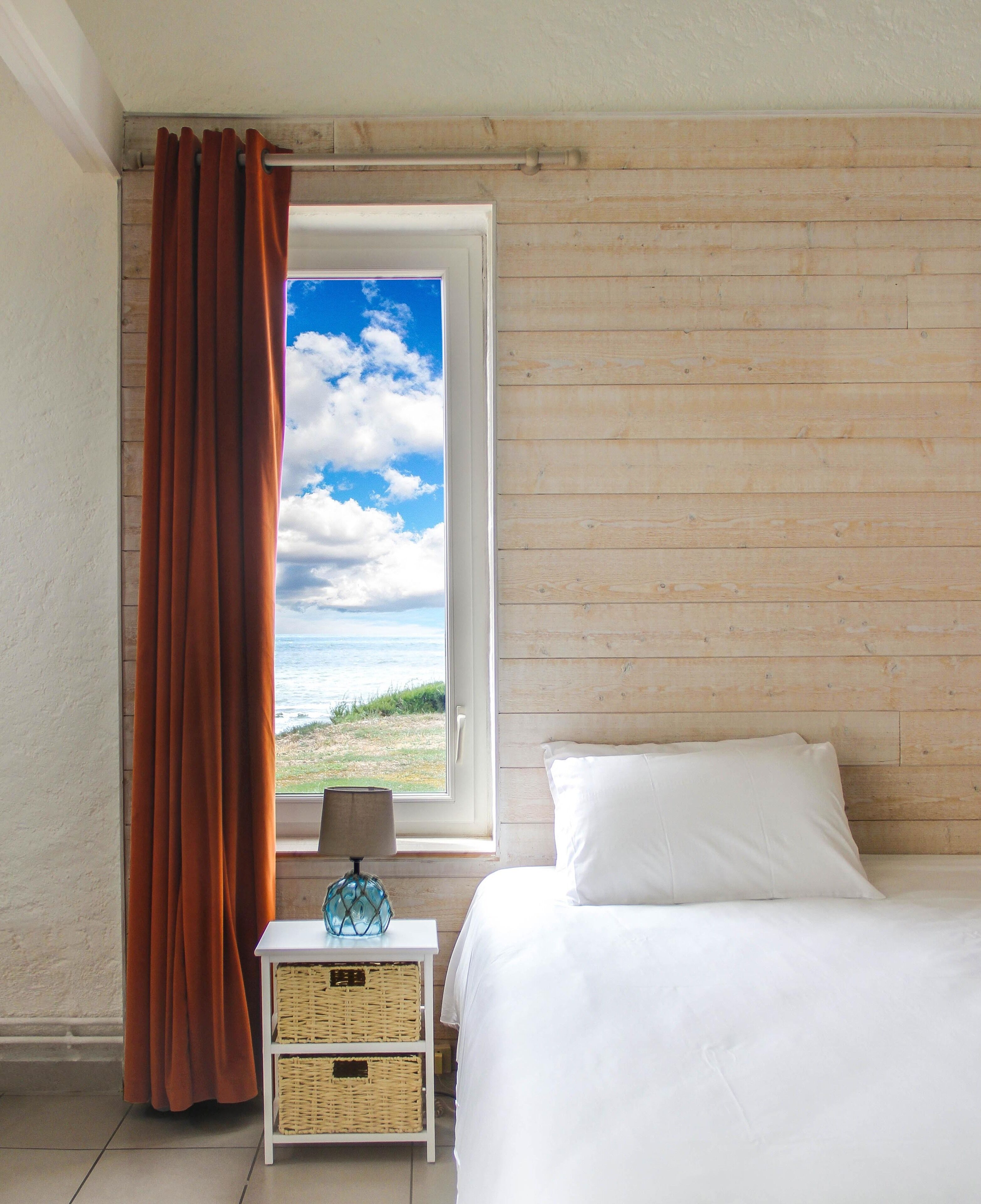 A bedroom with a wooden wall, a bed, a nightstand with a lamp, and a window with curtains.