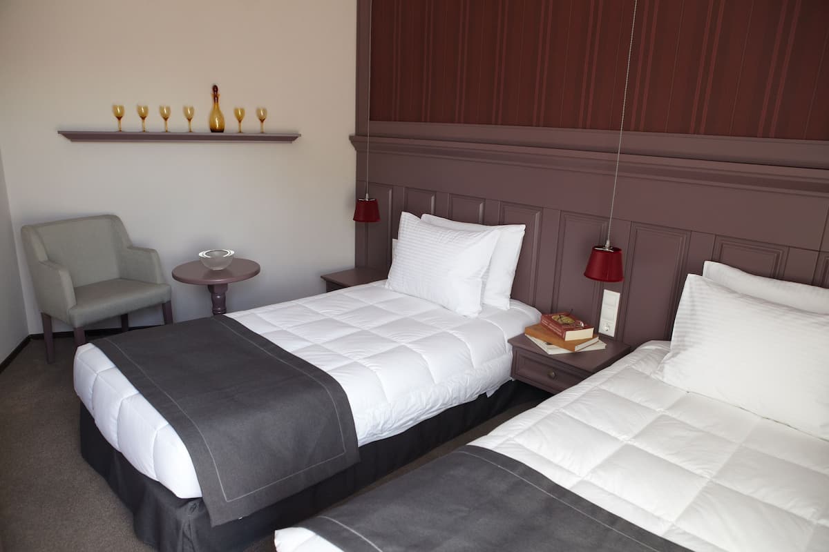 A hotel room with two beds, a chair, a small table, and a shelf with glasses and a bottle.