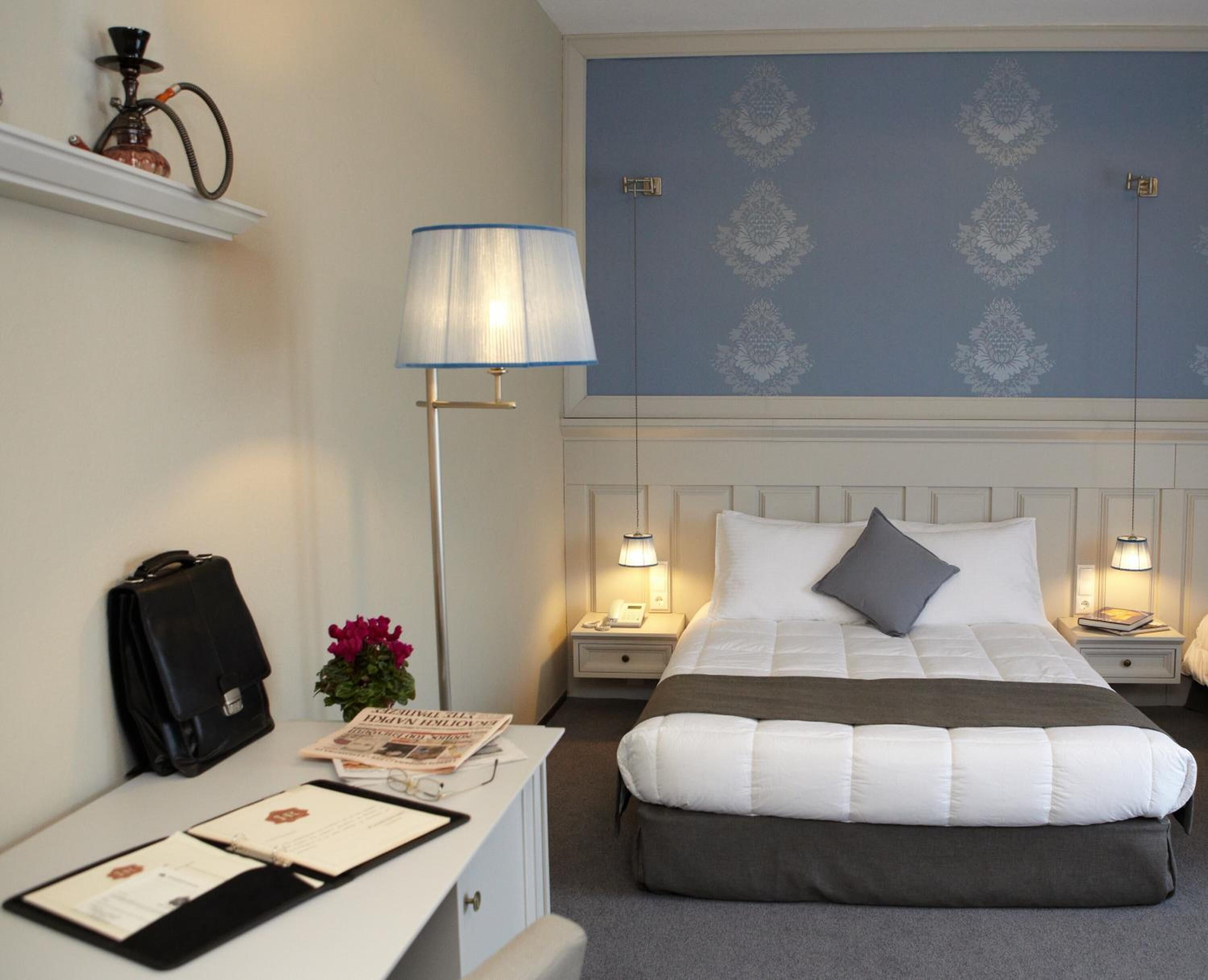 A hotel room with a bed, bedside tables, a desk, a lamp, a wall clock, and a decorative wall panel.