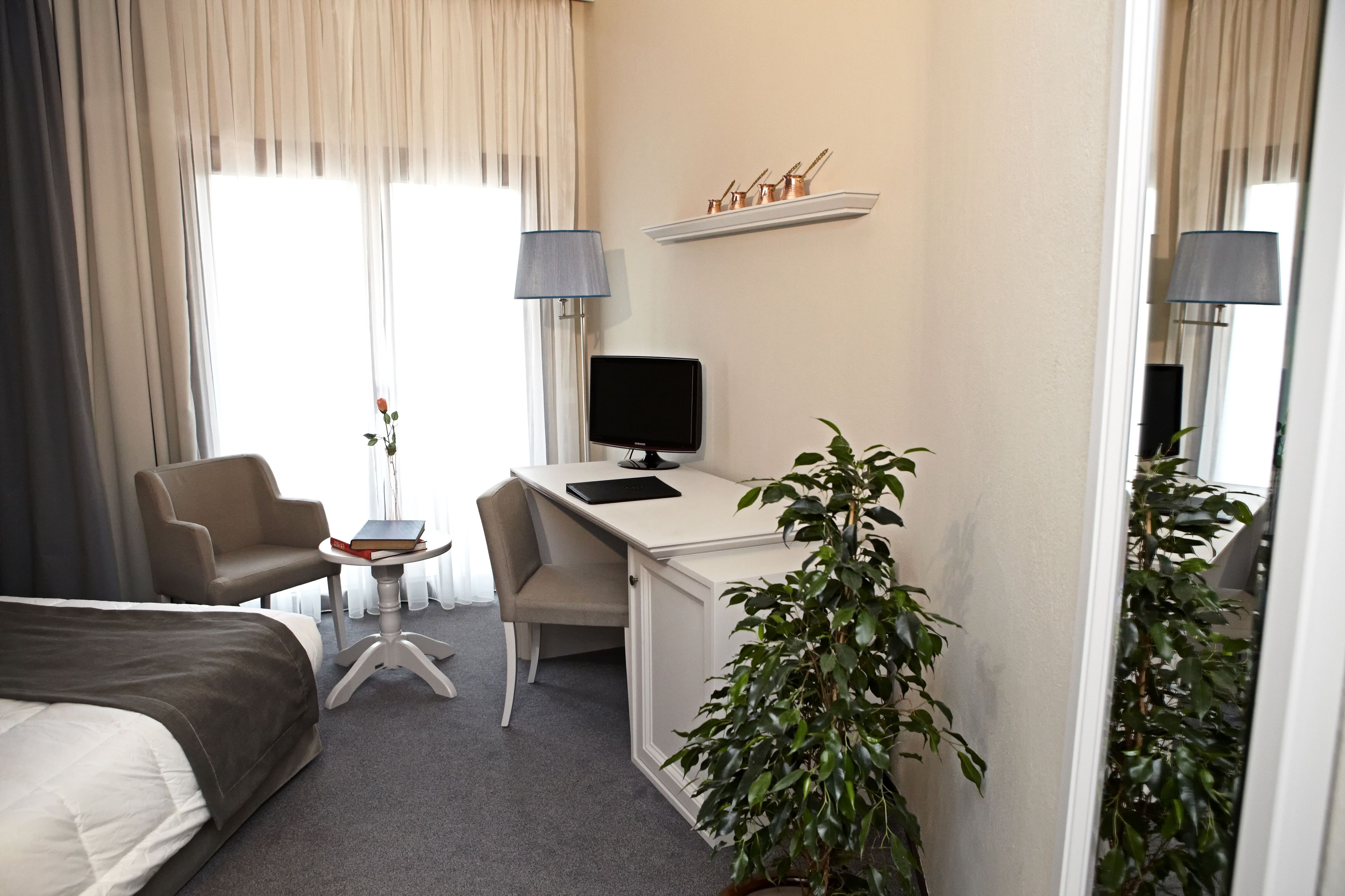A hotel room with a bed, a desk, a chair, a TV, a small table with a book and a vase, and a potted plant.
