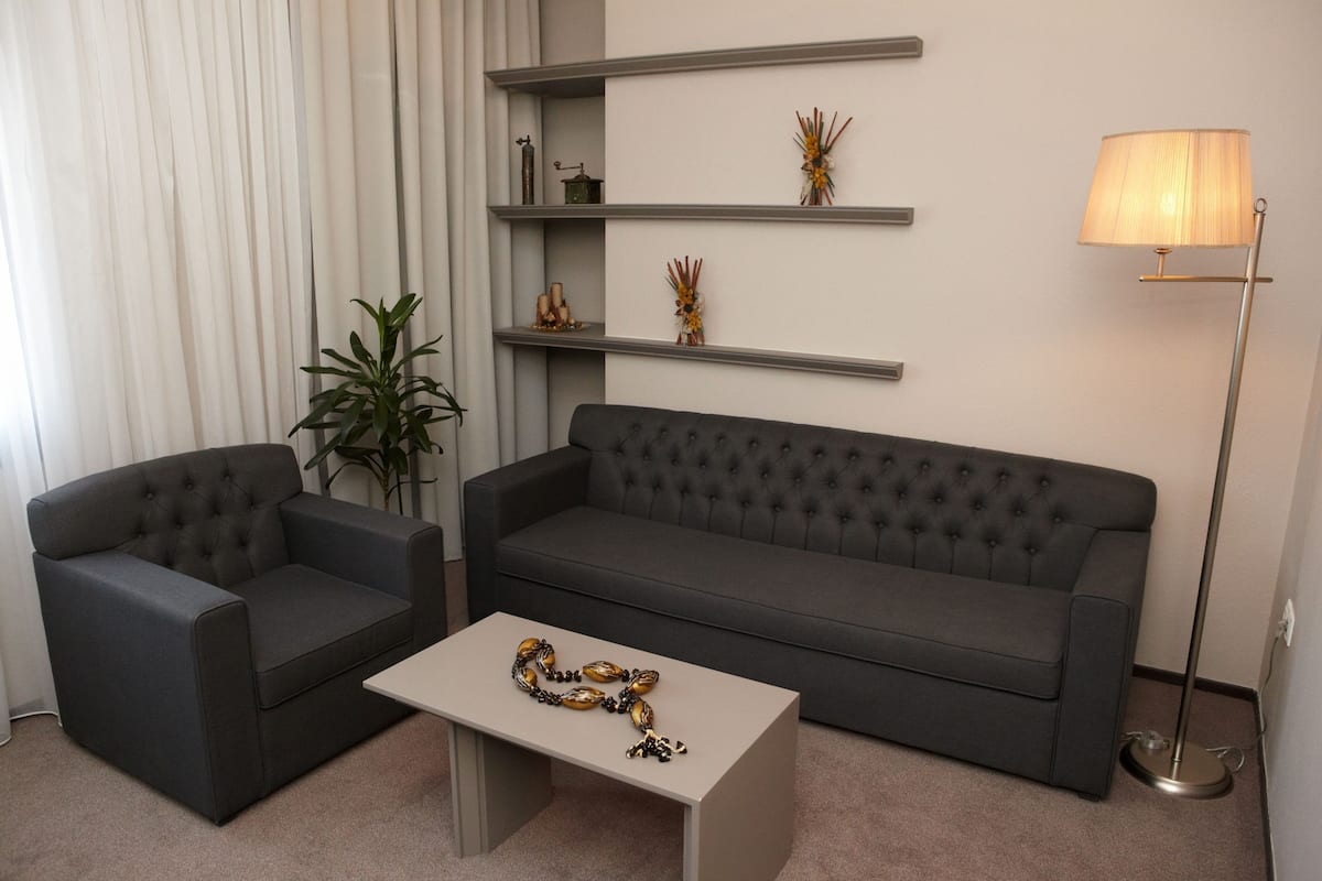 A modern living room with a grey sofa, a matching armchair, a coffee table, and a floor lamp. Shelves with decorative items are mounted on the wall.