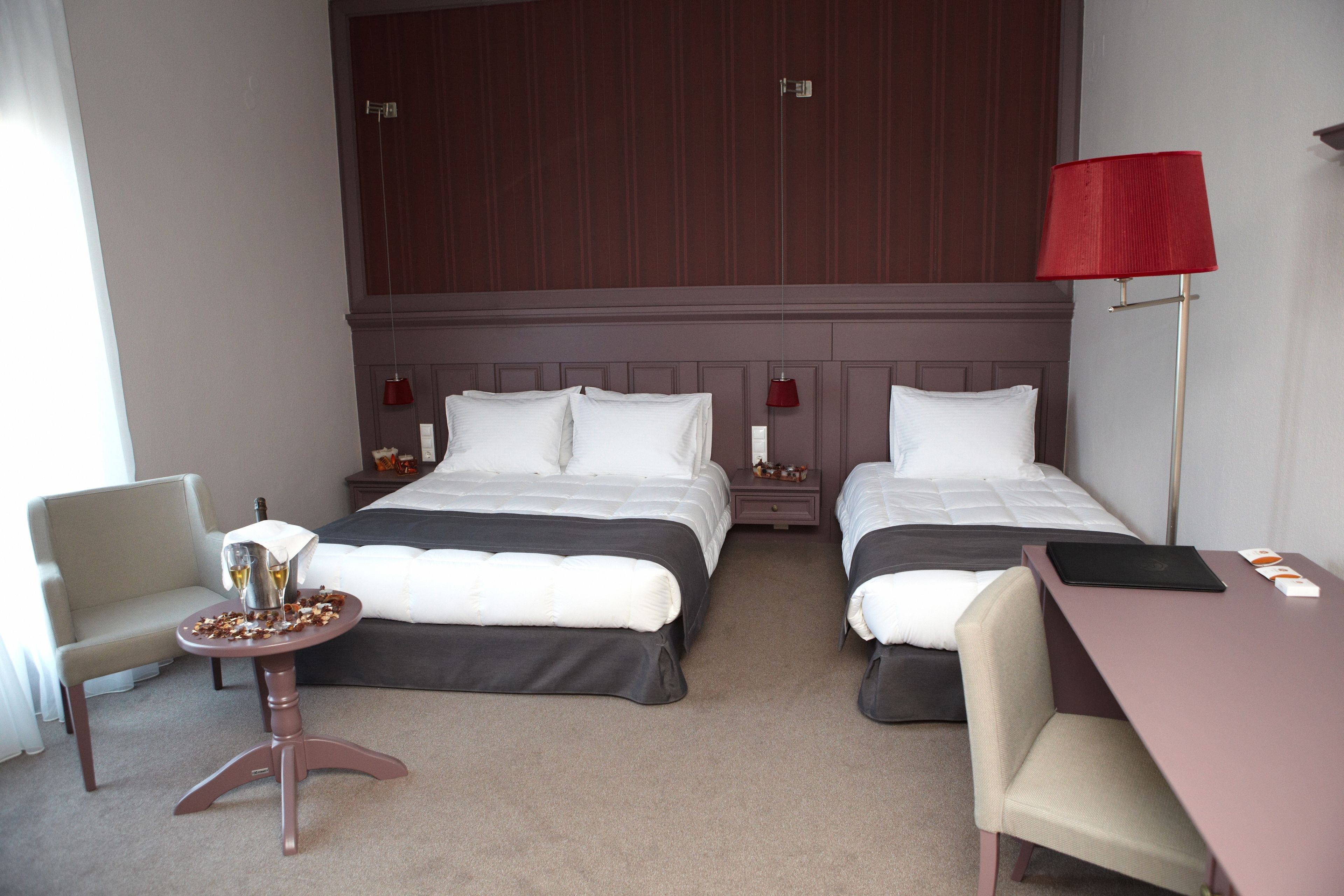 A hotel room with two beds, a desk, a chair, a small table, and a red lamp.