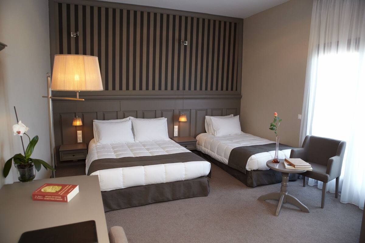 A hotel room with two beds, a desk, a chair, a lamp, and a small table with a book and a vase.