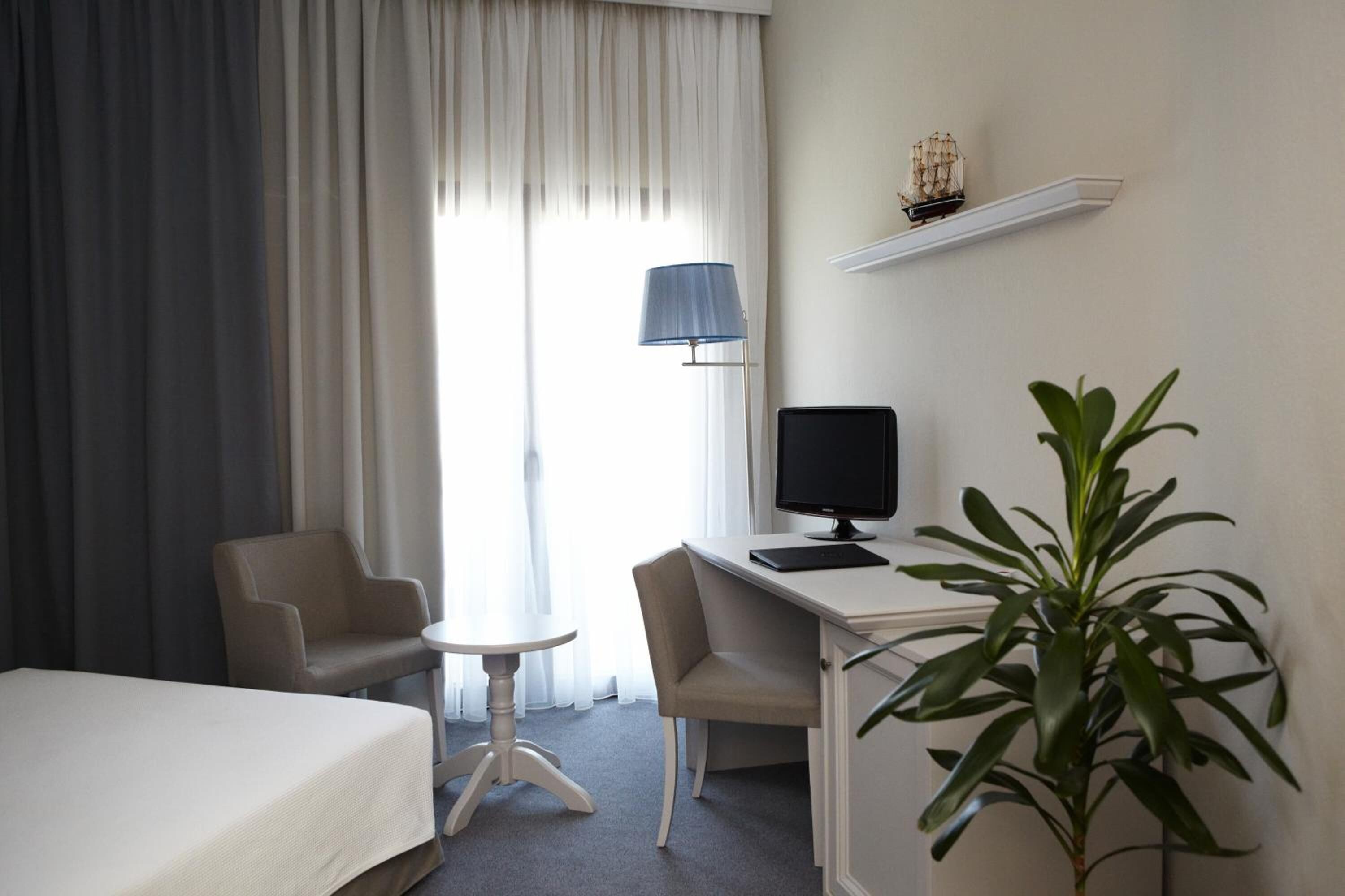 A hotel room with a bed, two chairs, a desk with a computer, a lamp, and a plant.
