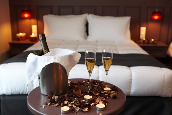 A neatly made bed with white linens, a bottle of champagne, two glasses of champagne, and scattered dried flowers on a bedside table.