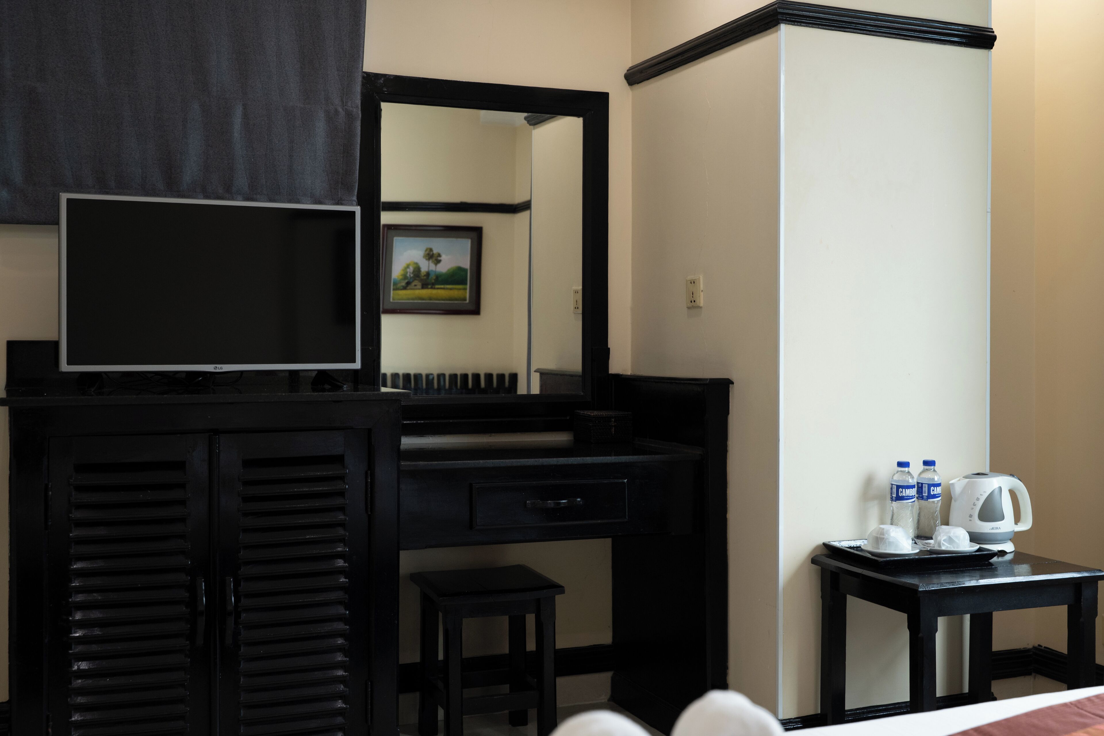 Habitación de hotel con un escritorio negro, televisor, taburete pequeño y bandeja con tetera y botellas de agua.