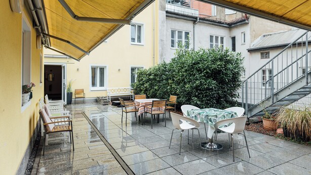 An outdoor patio with a yellow canopy, tables, chairs, and a large bush. - Hotel Andra (Munich)