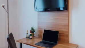 A wooden desk with a laptop, a bottle of champagne, and two glasses. A flat-screen TV is mounted above the desk. - Hotel Andra (Munich)