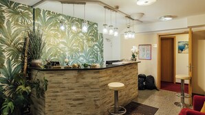 A reception area with a stone counter, two bar stools, a potted plant, and a tropical wallpaper. - Hotel Andra (Munich)