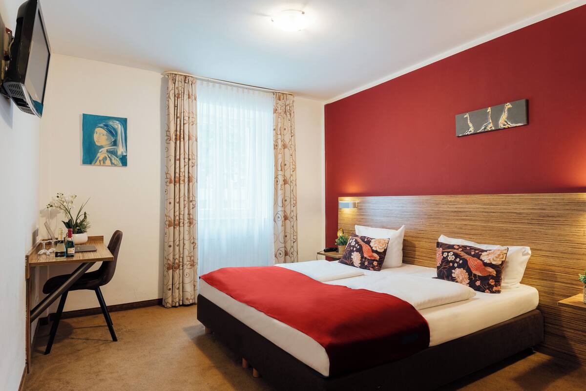 Hotellihuone, jossa on sänky, työpöytä, tuoli, televisio ja ikkuna, jossa on verhot.