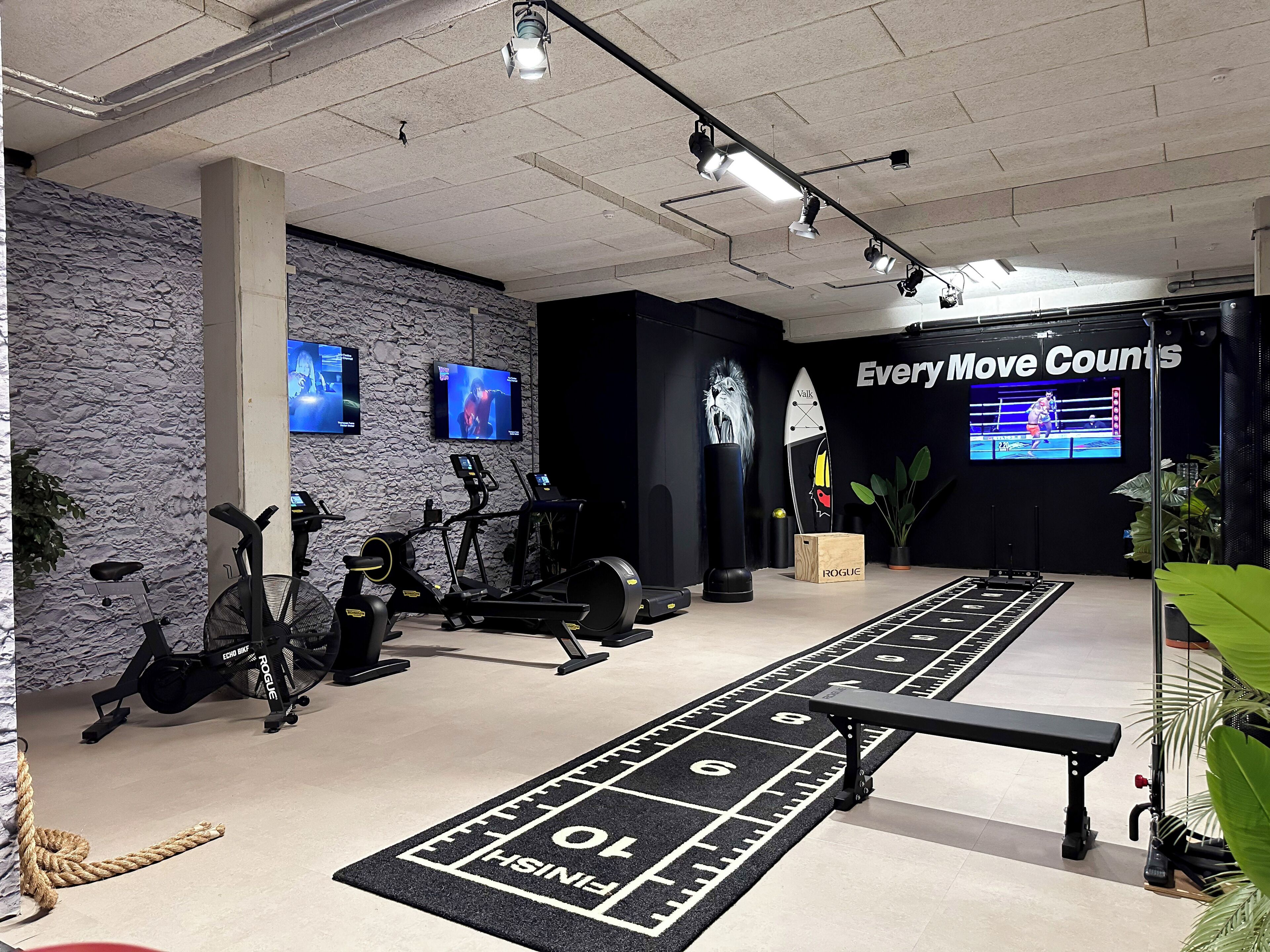 A gym with exercise equipment, a shuffleboard table, and a wall with a television displaying a boxing match.