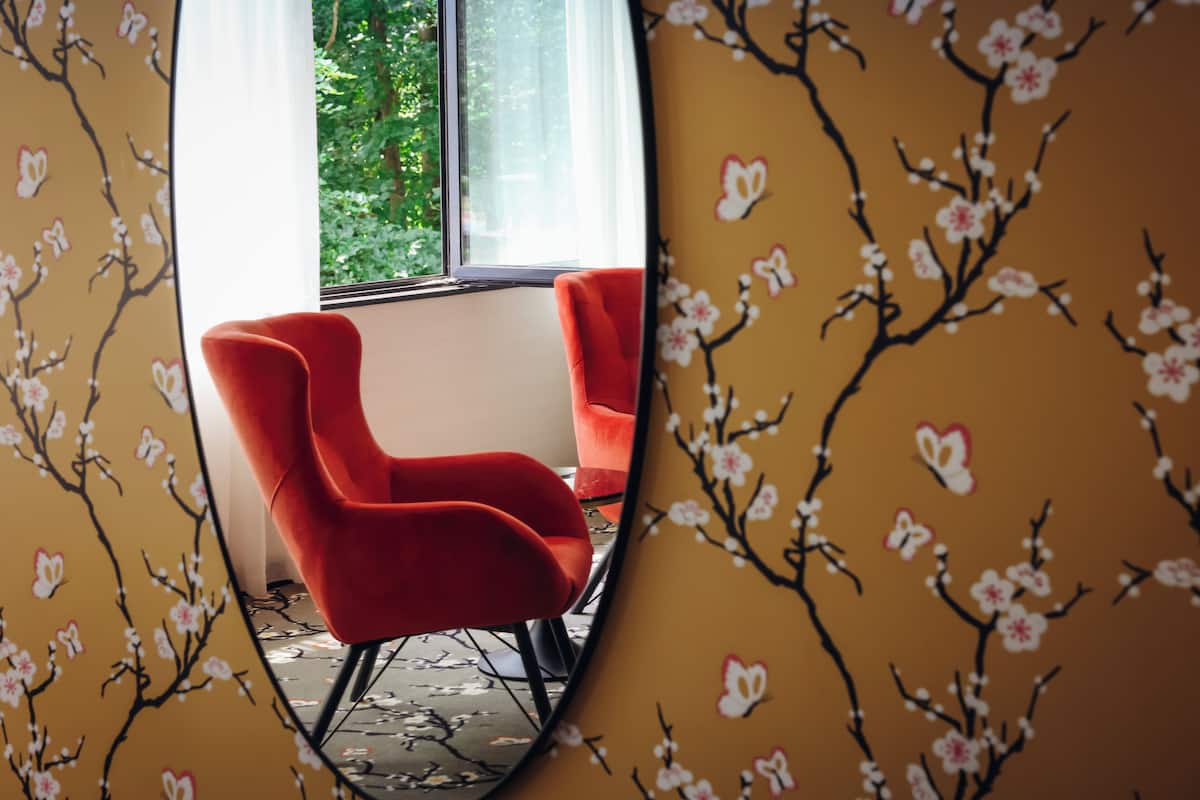 Ein Zimmer mit einem roten Sessel, einem fenster mit Vorhängen und einer Wand mit floralem Muster.