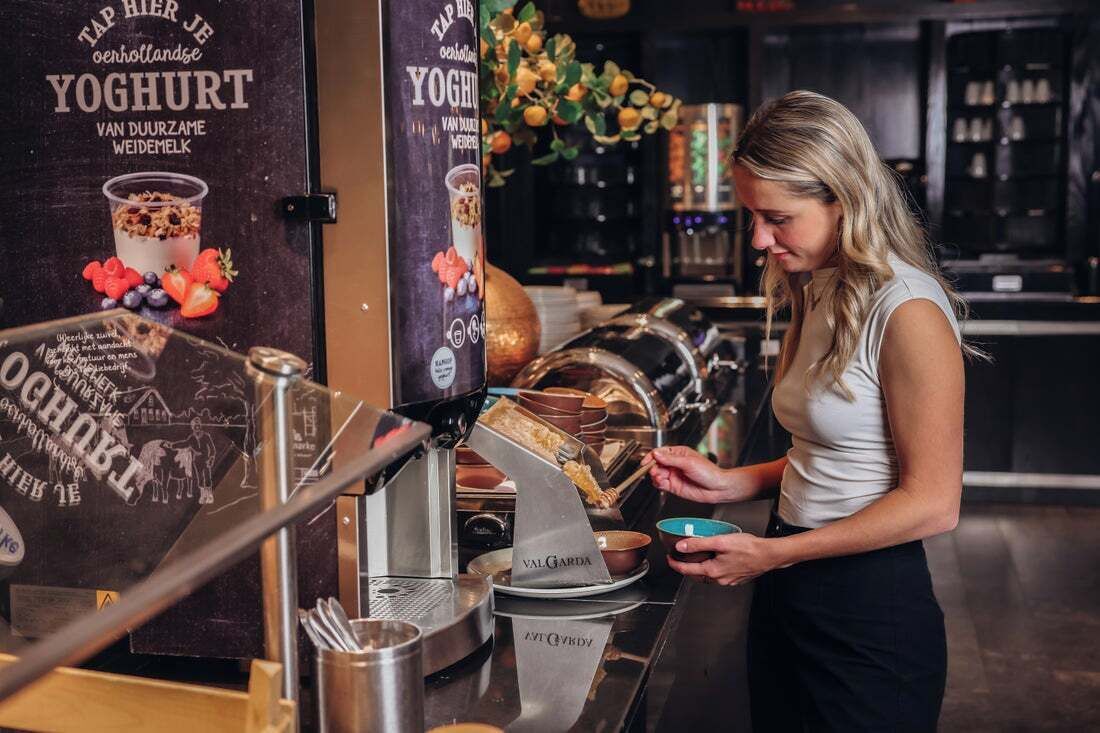Una persona preparando una bebida en un bar, con un anuncio de yogur y una máquina de café.