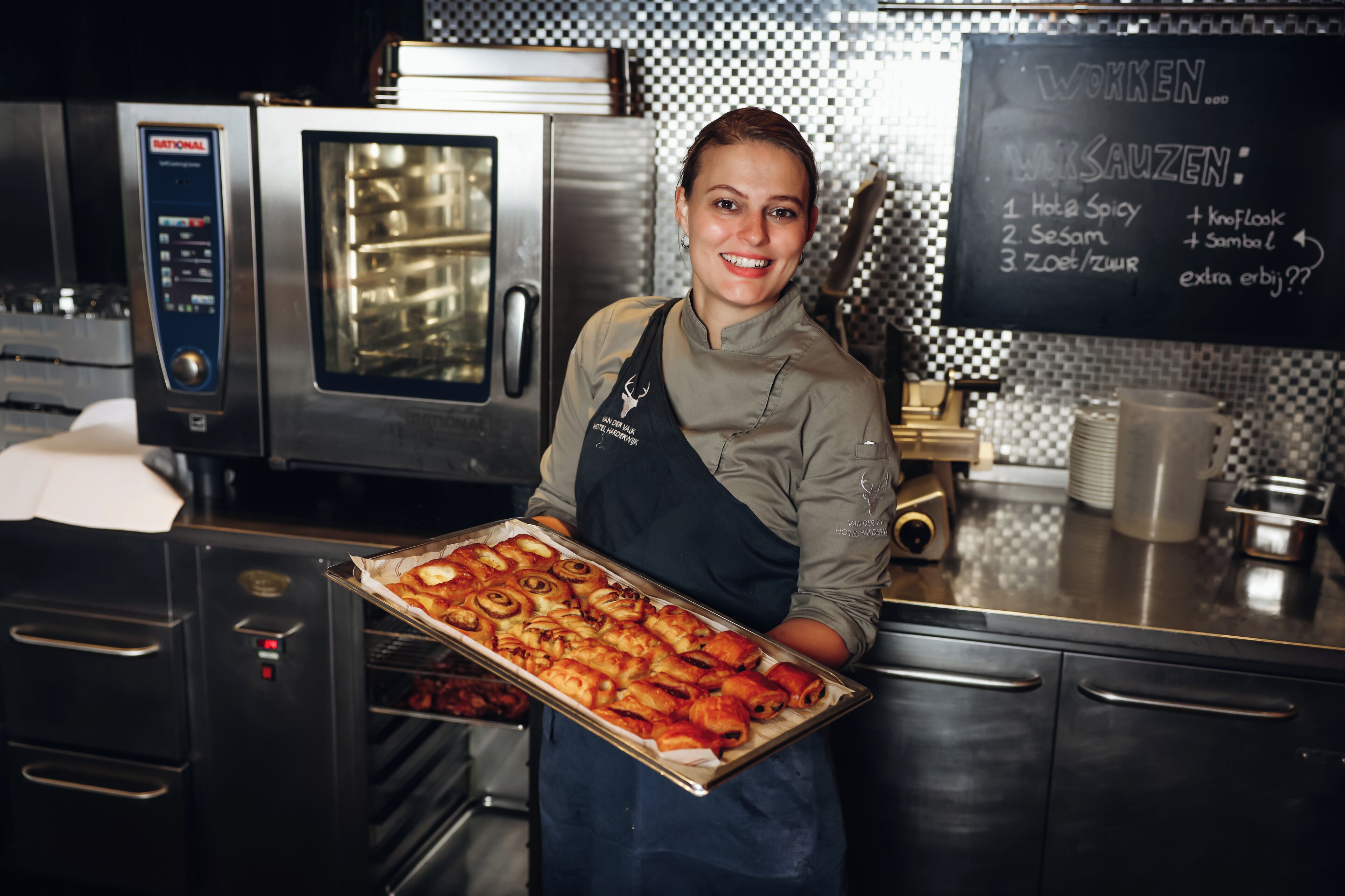 Een keuken met roestvrijstalen apparatuur, een krijtbordmenu en een persoon die een schaal gebak vasthoudt.