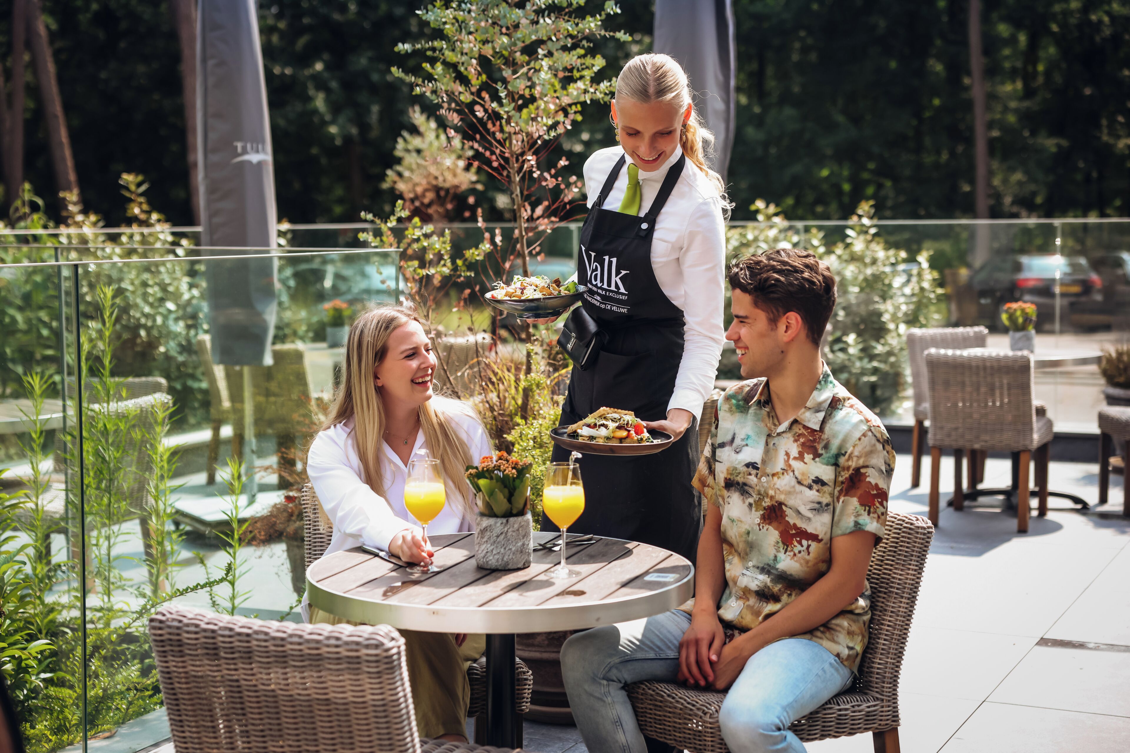 Un espace extérieur aménagé pour les repas, avec des meubles en osier, une table ronde et un serveur qui apporte les plats.