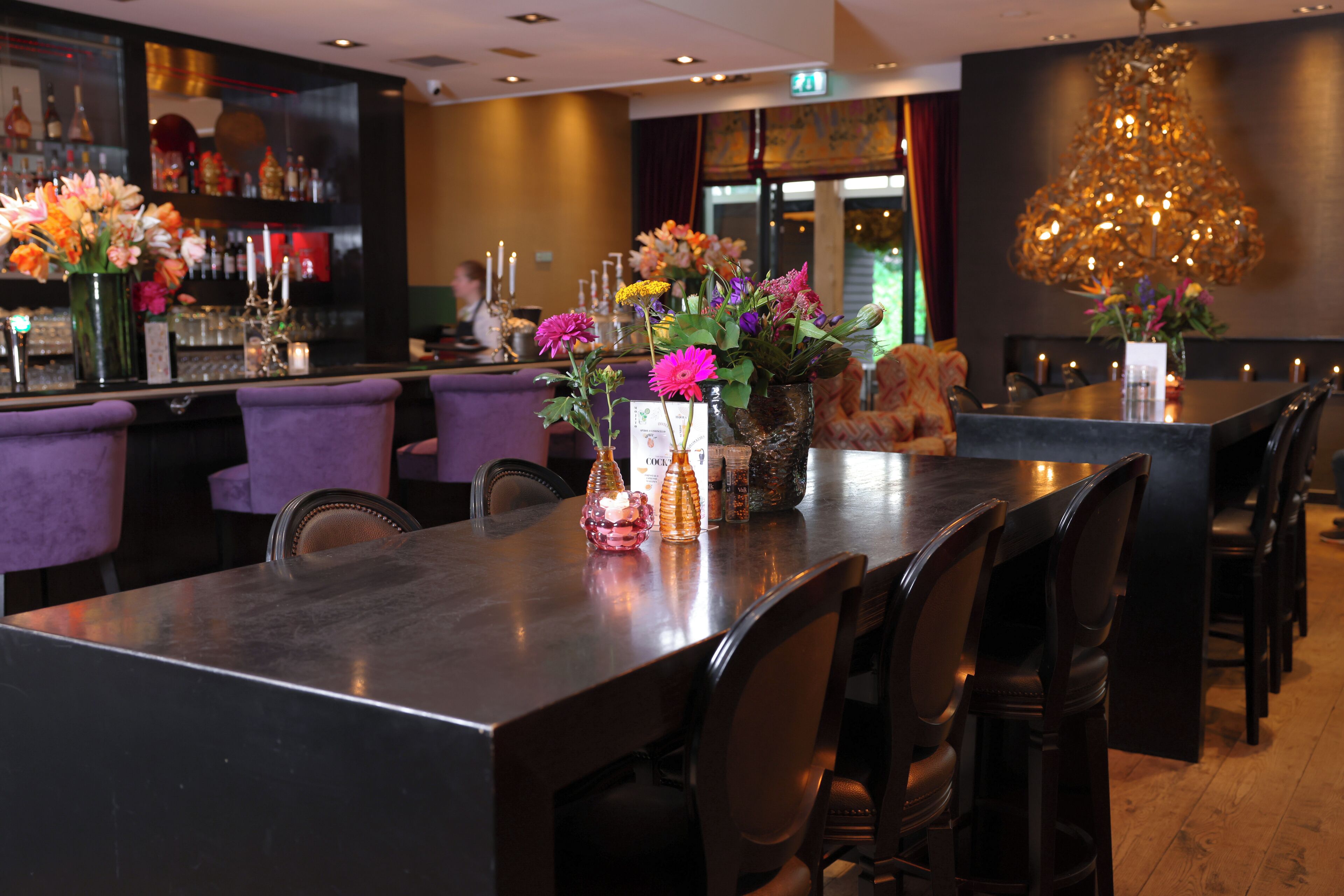 Un restaurante con una larga mesa, sillas moradas y una zona de bar con botellas y flores.