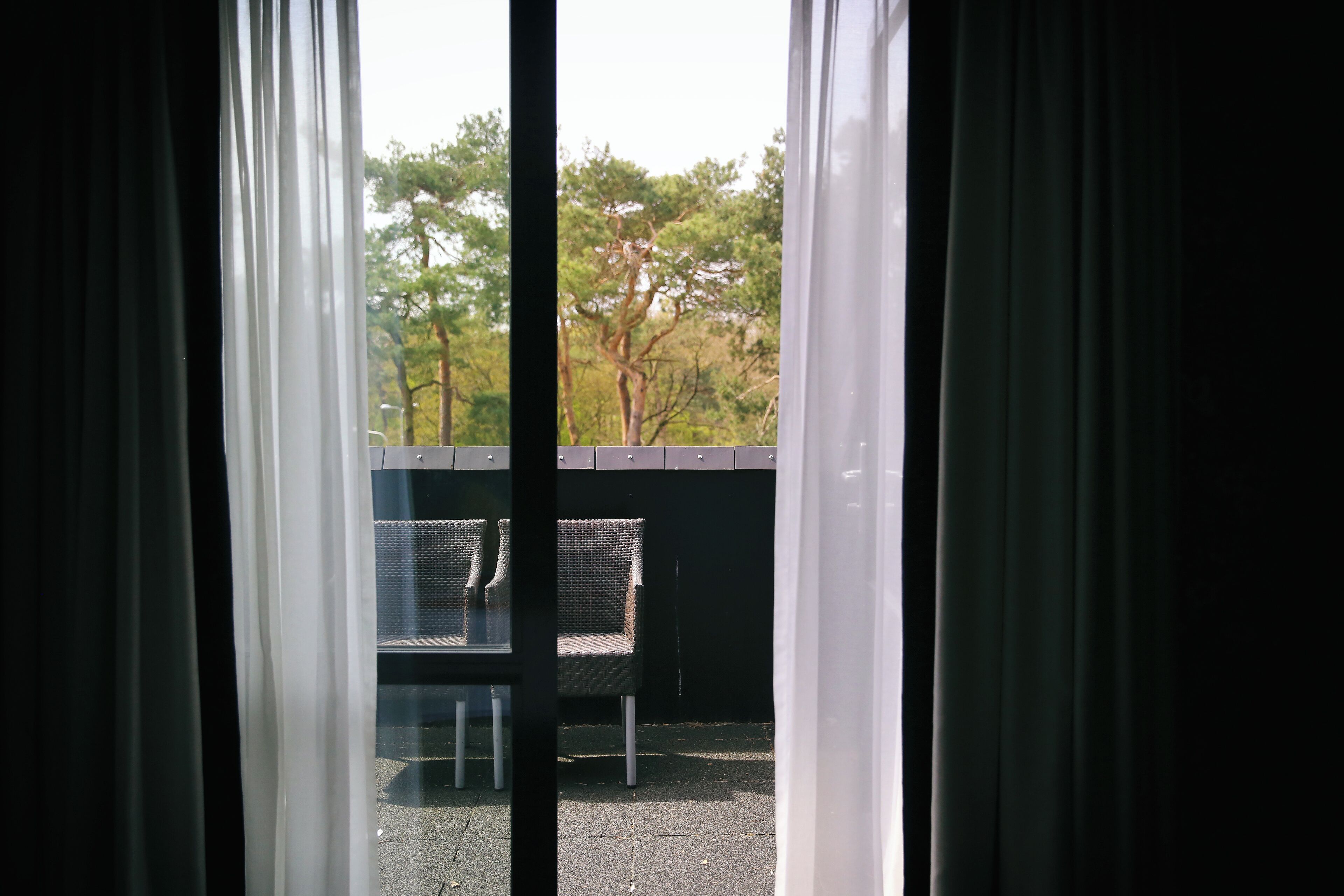 Vista pela janela com cortinas, mostrando área externa com mesas e cadeiras e árvores.