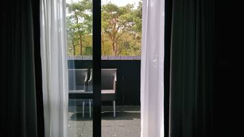 Vista pela janela com cortinas, mostrando área externa com mesas e cadeiras e árvores.