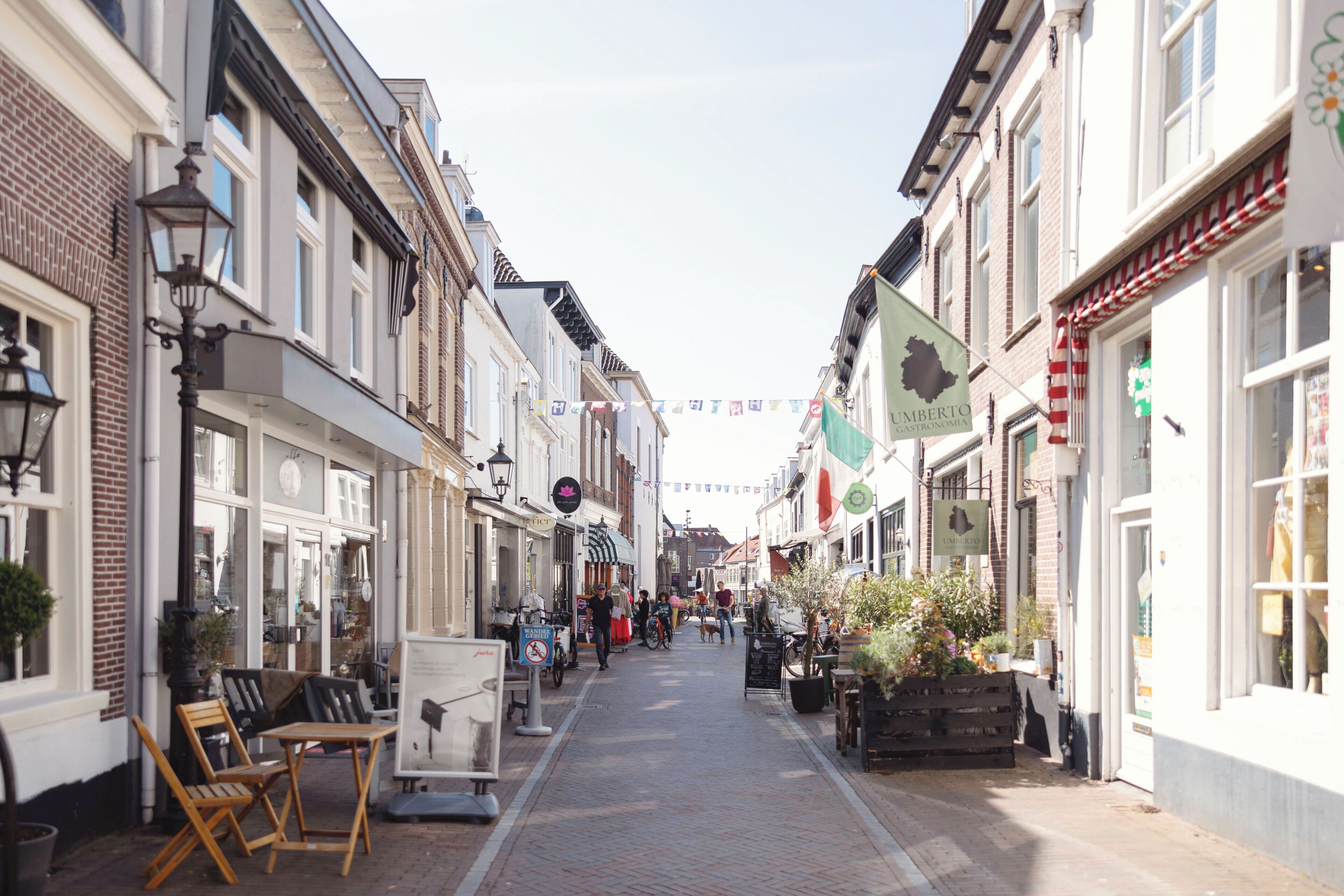A narrow street lined with shops and cafes, featuring outdoor seating and potted plants.