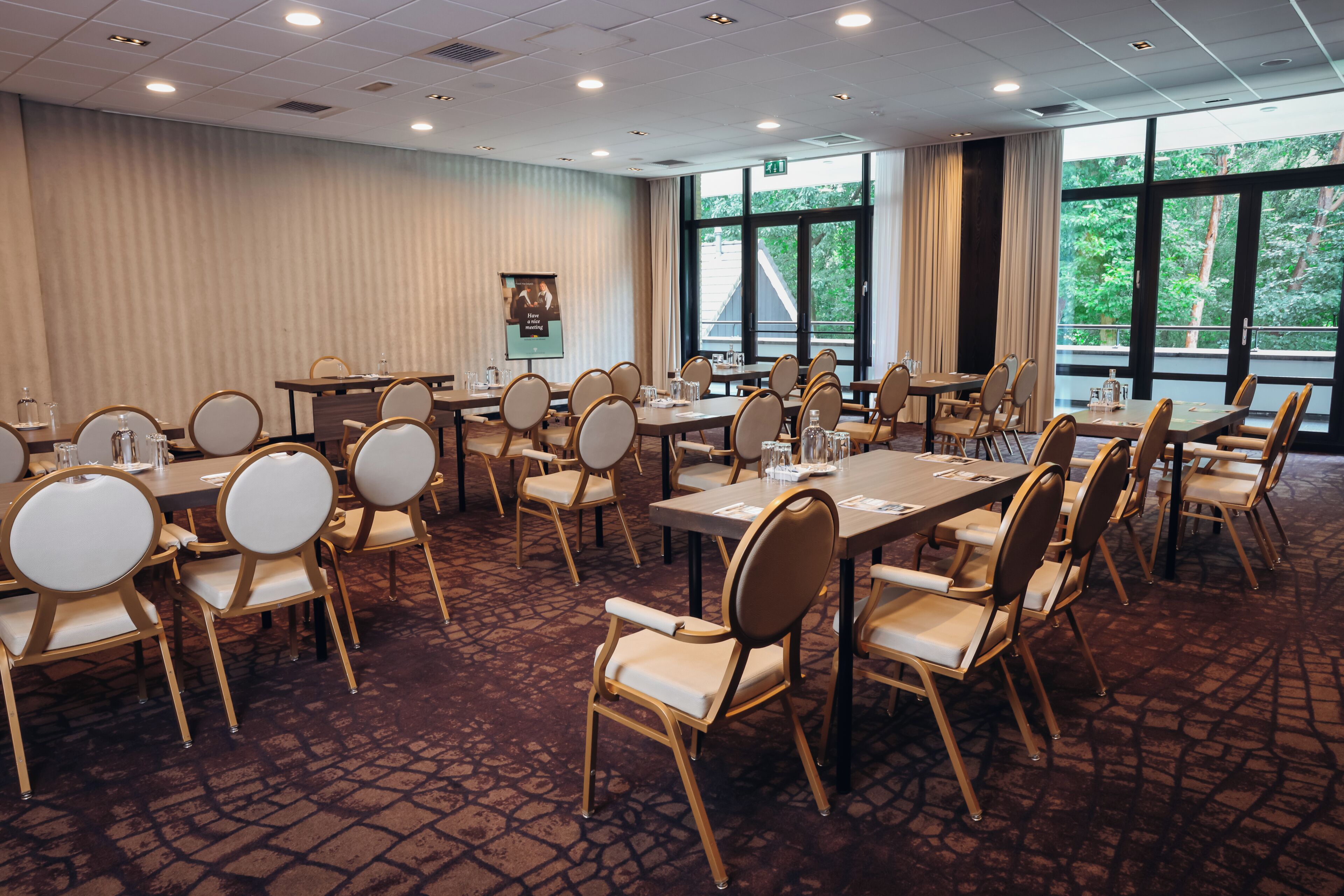 Un salón de conferencias con varias mesas y sillas, amplias ventanas con vistas a árboles y una alfombra estampada.