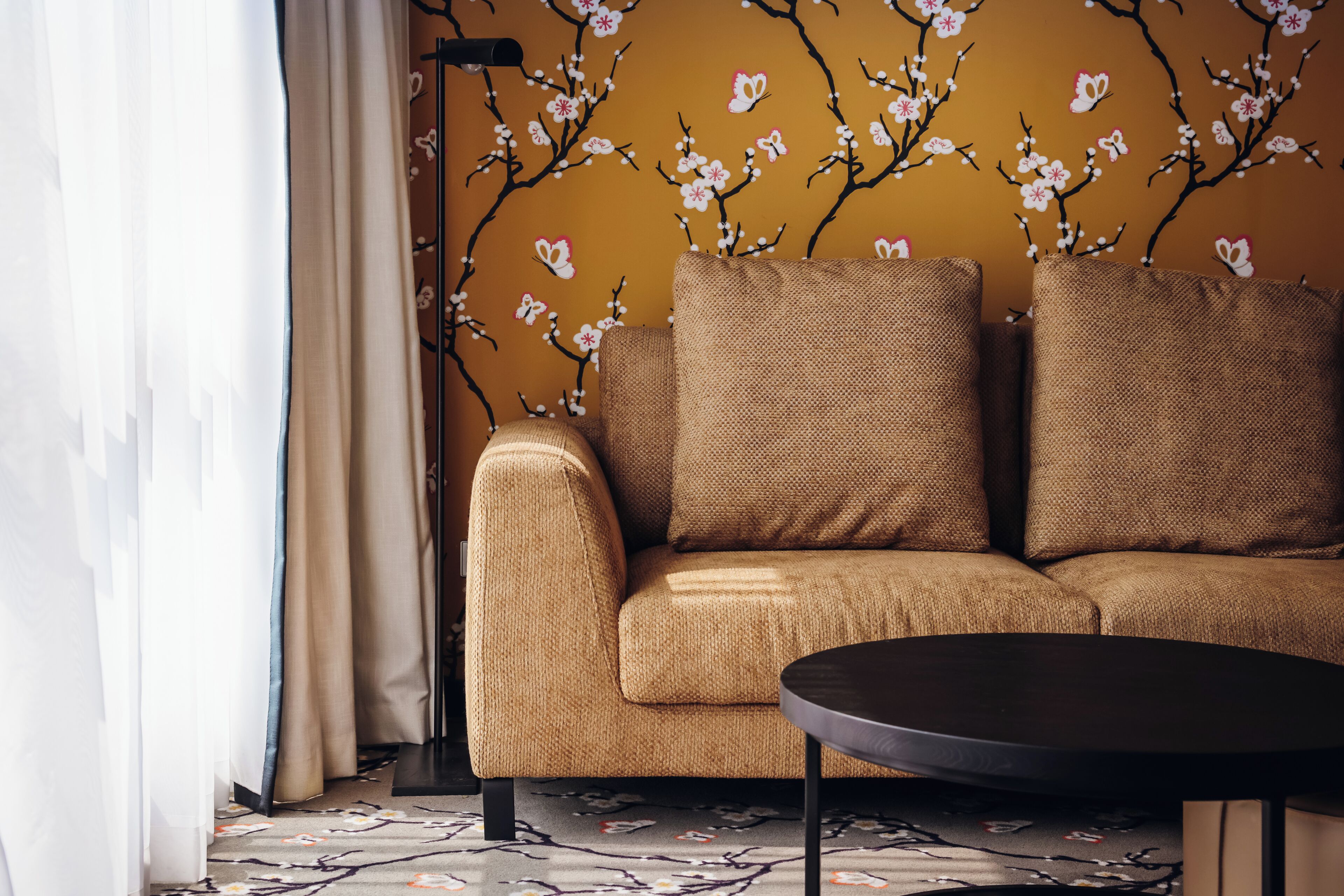 A sofa with a textured fabric, a round table, and a floor lamp in a room with a floral wallpaper.