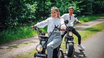 Twee personen rijden op elektrische scooters over een verhard pad, omringd door groen.