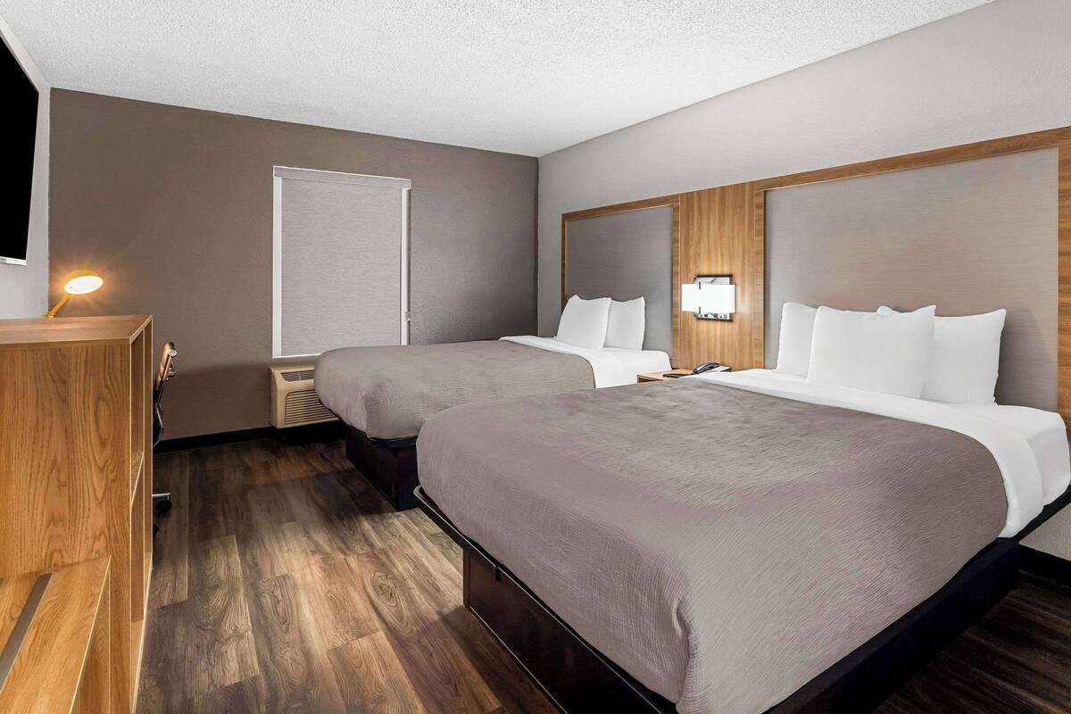 A hotel room with two beds, a wooden desk, a desk lamp, a wall-mounted TV, and a window with blinds.