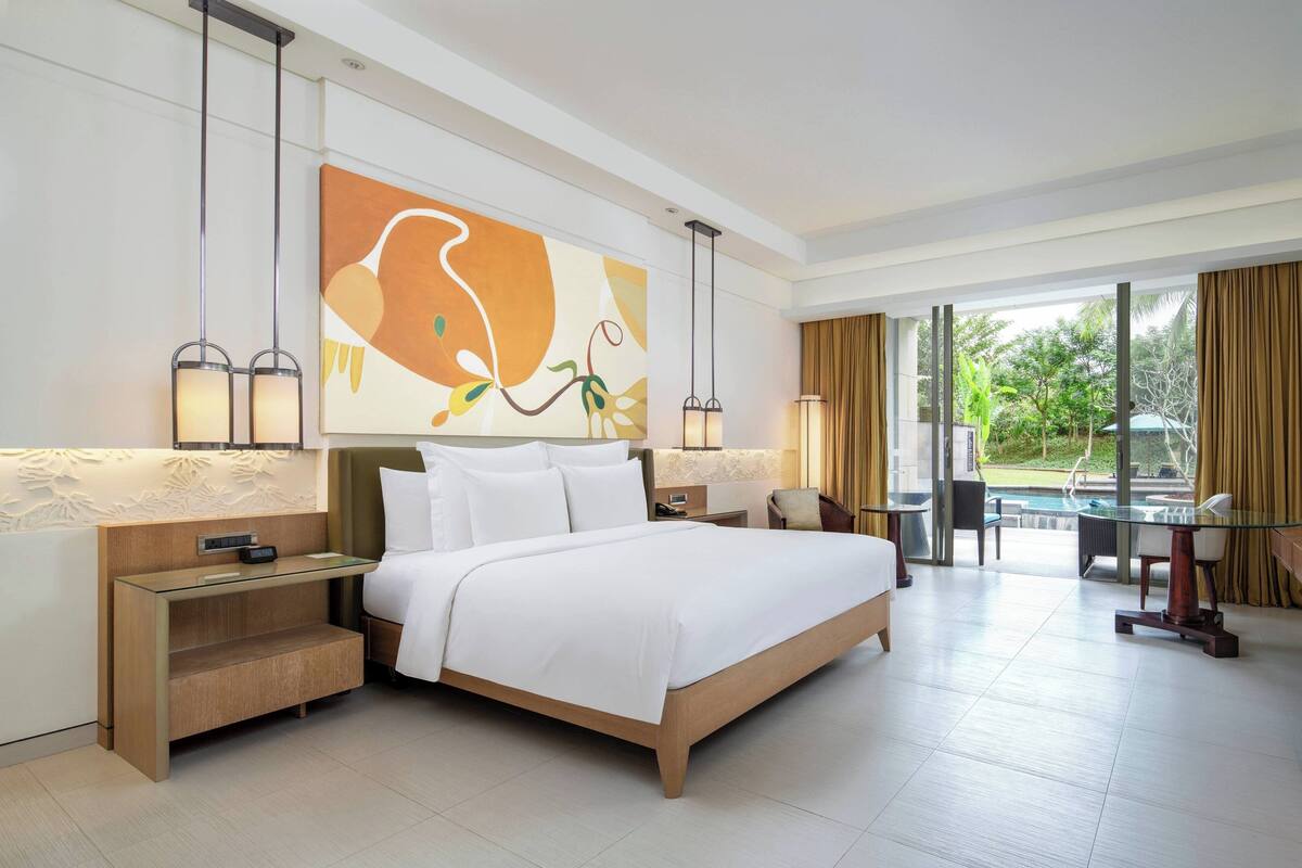 A modern hotel room with a large bed, bedside tables, a glass table, and a view of the outdoors.