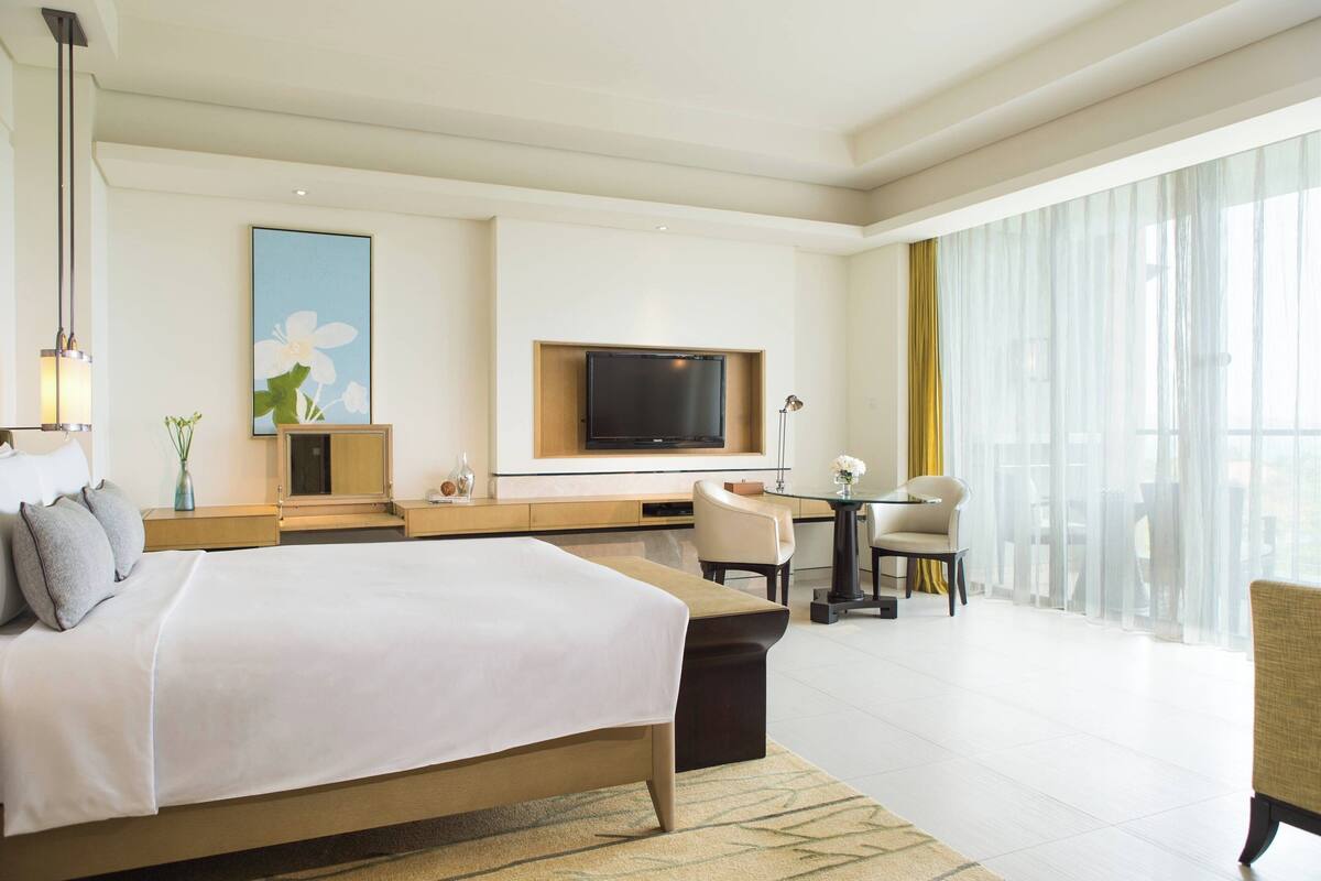 A modern hotel room with a large bed, a flat-screen TV mounted on the wall, a desk with a chair, and a large window with sheer curtains.