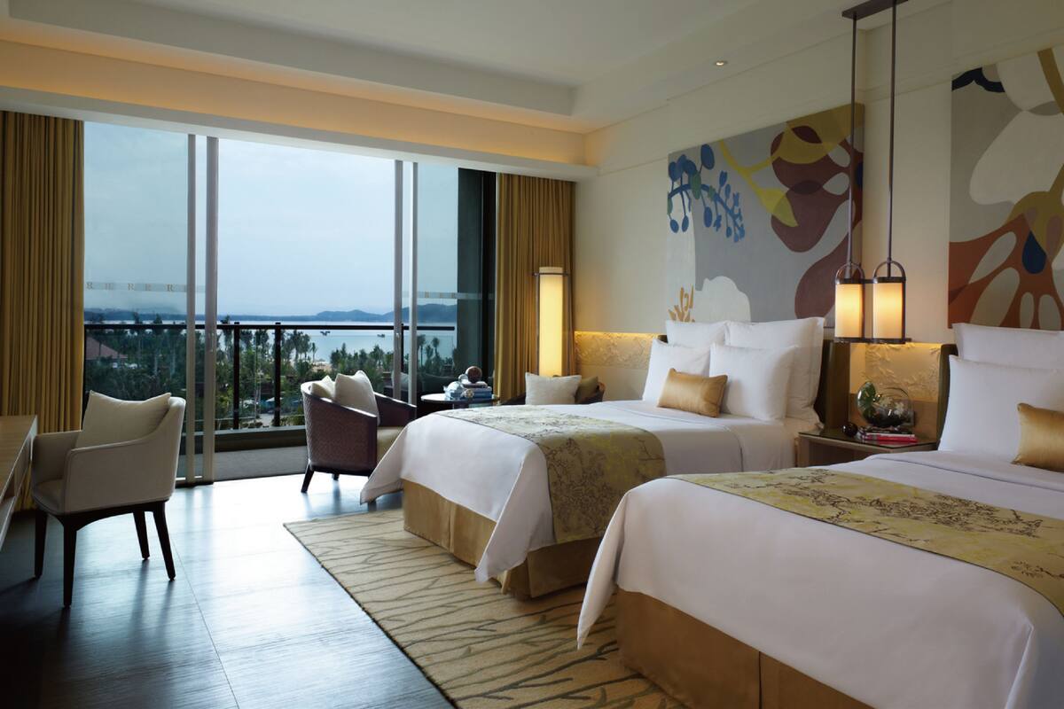 A hotel room with two beds, a sitting area with a chair, a large window with a view, and a decorative wall piece.
