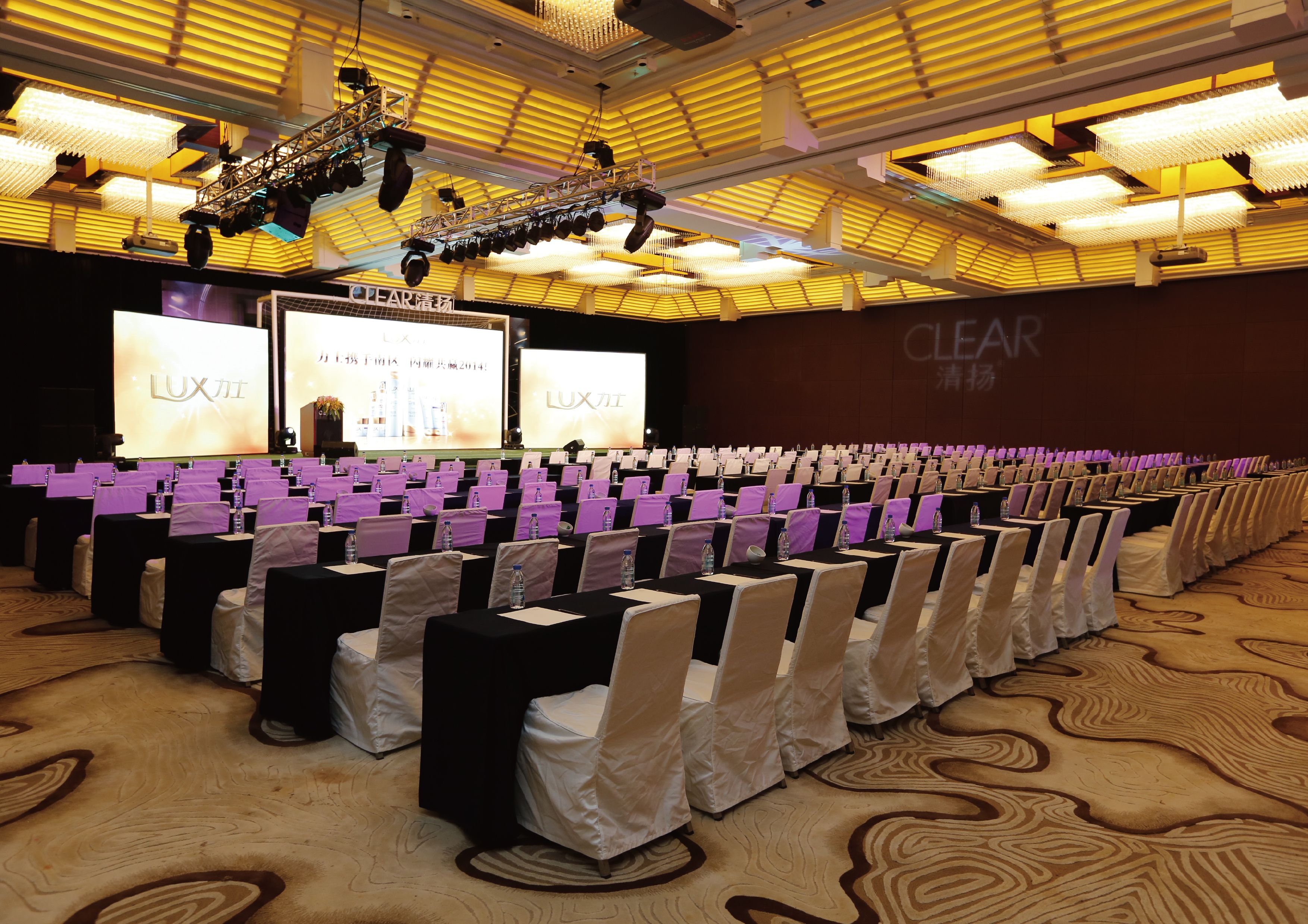 A conference room with rows of tables and chairs, a stage with a large screen, and a ceiling with patterned lights.